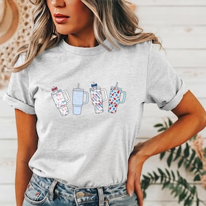 May include: A light gray short-sleeve t-shirt featuring a graphic print of four insulated tumblers with straws. The tumblers are decorated with various red, white, and blue patterns, including small bows, stars, and stripes, arranged in a horizontal row.