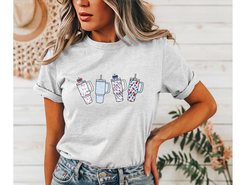 May include: A light gray short-sleeve t-shirt featuring a graphic print of four insulated tumblers with straws. The tumblers are decorated with various red, white, and blue patterns, including small bows, stars, and stripes, arranged in a horizontal row.