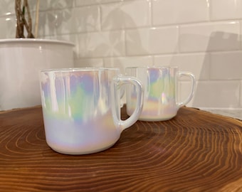 May include: Two iridescent white ceramic mugs with handles on a wooden surface.