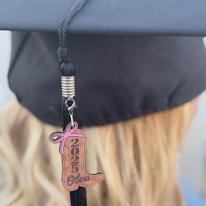 Op de afbeelding: Een zwarte afstudeerhoed met een kwast. Een houten bedel in de vorm van een cowboylaars is aan de kwast bevestigd. De bedel heeft het jaar 2025 en de naam Olivia, met een roze lint.