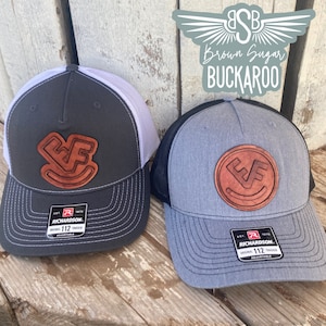 May include: Two trucker hats with leather patches. One is dark gray with a white mesh back, the other is gray with a black mesh back. Both have a brown leather patch with the letters "EF" and a smiling face. The hats are labeled "Richardson 112 Trucker".