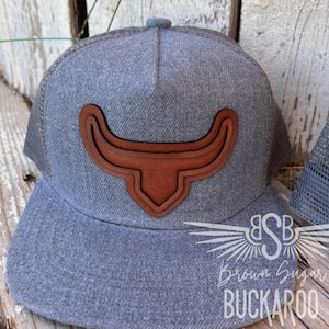 May include: Gray and brown trucker hat with a brown leather bull horn design. The hat has a mesh back and the words "Brown Sugar Buckaroo" are embroidered on the front.