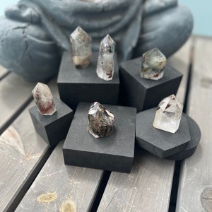 Könnte beinhalten: Klare Quarz-Kristallspitzen mit Einschlüssen, präsentiert auf schwarzen geometrischen Podesten. Im Hintergrund ein großer, rauer Amethystkristall, mit einer grauen Statue und Holzplanken.