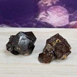 Könnte beinhalten: Zwei dunkelbraune und schwarze Granat-Kristalle mit einer rauen, facettierten Textur. Die Kristalle liegen auf einer weißen Holzoberfläche.