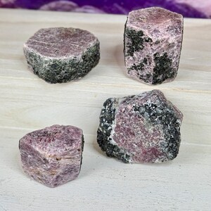 Könnte beinhalten: Vier rosafarbene Rubinkristalle mit schwarzen Einschlüssen. Die Kristalle sind roh und haben ein natürliches, unpoliertes Aussehen.