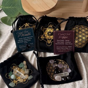 Könnte beinhalten: Mehrere schwarze Samt-Kordelzugbeutel mit goldenen geometrischen Designs und kleinen Kristallen im Inneren. Jeder Beutel hat eine Karte mit dem Text: "Crystal Grid for Wealth" oder "Crystal Grid for Love". Die Kristalle sind verschiedenfarbig.