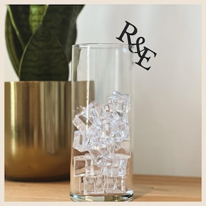 May include: A tall, clear glass filled with ice cubes, with the letters "R&E" on a black tag. A gold-colored pot with a green plant is in the background. The glass is on a wooden surface.