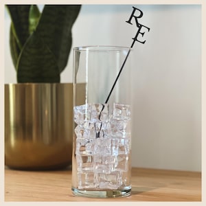 May include: A clear, cylindrical glass filled with ice cubes. A black cocktail pick with the letters "RE" is placed inside the glass. A gold-colored pot with a green plant is in the background.