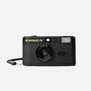 May include: Black ANIKEN camera with a gold logo, a flash, and a lens that reads "FOCUS FREE OPTICS LENS 28MM F/8". The camera has a wrist strap attached. The camera is a compact, point-and-shoot style.