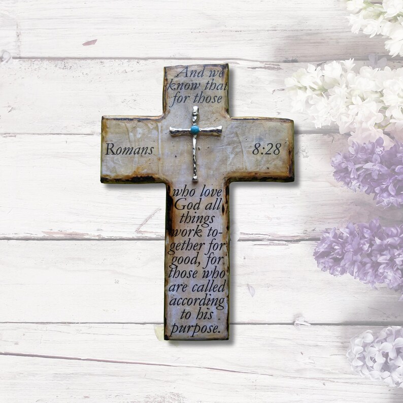 May include: A rustic wooden cross with a distressed white finish and a silver cross with a turquoise stone. The cross features the Bible verse Romans 8:28, which reads "And we know that for those who love God all things work together for good, for those who are called according to his purpose."