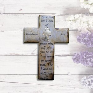 May include: A rustic wooden cross with a white background and a silver cross charm. The cross features the text "Now may the Lord of peace himself give you peace at all times in every way. The Lord be with you all." and the scripture reference "2 Thessalonians 3:16".