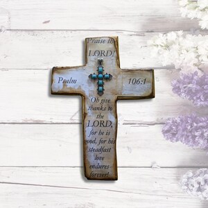 May include: A wooden cross with a distressed white paint finish and a turquoise and silver cross charm. The cross has the text "Psalm 106:1" and "Praise the LORD!"  "Oh give thanks to the LORD, for he is good, for his steadfast love endures forever!"