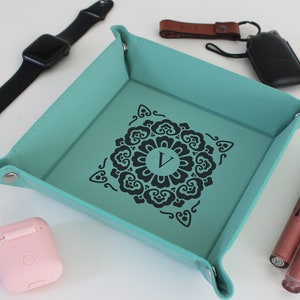 Könnte beinhalten: Ein tealfarbenes Leder-Ablagefach mit einem schwarzen Mandala-Design und dem Buchstaben "V" in der Mitte. Das Fach befindet sich auf einer weißen Oberfläche mit einer schwarzen Uhr, einer rosa Handyhülle, einem braunen Leder-Schlüsselanhänger, einer schwarzen Handyhülle und zwei Lippenstift-Tuben.