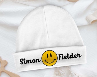 Pode incluir: Gorrinho branco para bebé com um sorriso amarelo e o texto "Simon Fielder" em preto.