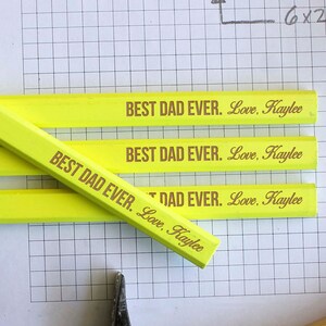 Pode incluir: Quatro lápis de carpinteiro amarelos com o texto "BEST DAD EVER. Love, Kaylee" impresso neles. Os lápis estão sobre uma folha de papel quadriculado branco com um martelo e parafusos no fundo.
