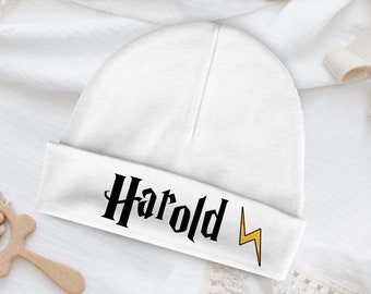 Pode incluir: Gorrinho branco para bebé com o nome "Harold" e um raio amarelo.