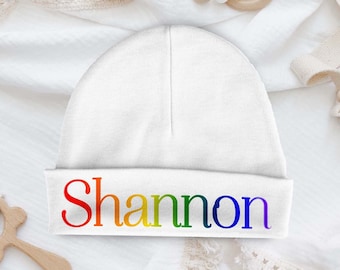 Op de afbeelding: Witte baby beanie met de naam "Shannon" in regenboogkleuren.