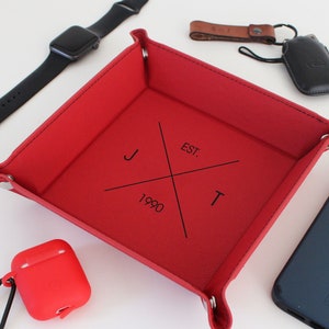 May include: Red leather valet tray with a black monogram that reads "J & T EST. 1990". The tray is filled with various items, including a black Apple Watch, a red AirPod case, a blue iPhone, and a black leather key fob.