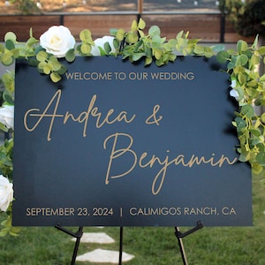 Puede incluir: Un letrero de bienvenida de boda negro con letras doradas. Dice "Bienvenida a nuestra boda" y presenta los nombres "Andrea & Benjamin". El letrero está adornado con rosas blancas y vegetación, exhibido en un caballete negro.