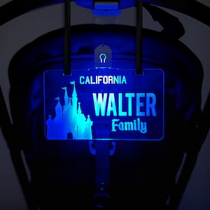Op de afbeelding: Een blauw verlicht kenteken met de tekst "CALIFORNIA" en "WALTER Family". Een kasteelsilhouet staat links. De plaat is bevestigd aan een zwarte kinderwagen.