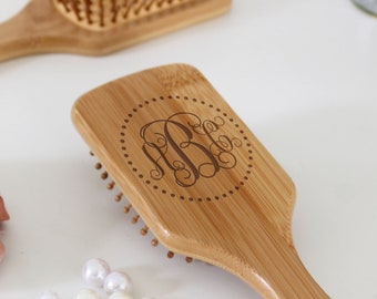 Puede incluir: Un cepillo de pelo de bambú con mango y cabeza rectangular. La cabeza presenta un diseño de monograma dentro de un círculo punteado. Varias perlas blancas y otro cepillo están en el fondo.