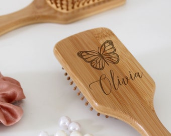 Puede incluir: Un cepillo para el cabello de madera con un acabado natural. El cepillo tiene una cabeza rectangular con un diseño de mariposa y el nombre "Olivia" grabado en él. El cepillo tiene un mango largo y redondeado.