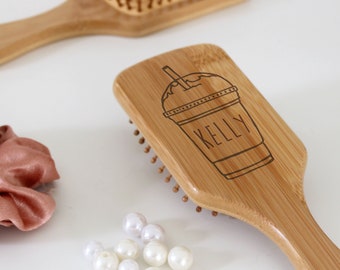 Puede incluir: Un cepillo para el cabello de madera con un mango marrón y cerdas naturales. El cepillo tiene un diseño personalizado con un dibujo de una taza y el nombre "Kelly" grabado en él.