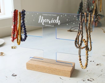Könnte beinhalten: Ein durchsichtiger Acryl-Schmuckständer mit Holzsockel. Der Ständer hat vier Fächer zum Aufbewahren von Halsketten und Armbändern. Das obere Fach trägt den Aufdruck "Things that make Mariah Sparkle".