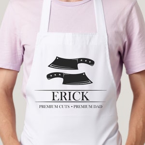 以下が含まれることがあります： 2つの黒い肉切り包丁のイラストが描かれた白いエプロン。「ERICK」という名前が太字で表示され、その下に「PREMIUM CUTS • PREMIUM DAD」というテキストがあります。エプロンには首ループとウエストタイが付いています。