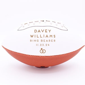 Pode incluir: Bola de futebol branca com costuras marrons e um painel de couro marrom. A bola é personalizada com o texto "Davey Williams Ring Bearer 11.23.24" e duas alianças douradas.
