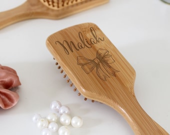 Puede incluir: Un cepillo para el cabello de madera con un acabado natural. El cepillo tiene una forma rectangular y un mango. El mango está grabado con el nombre "Maliah" y un lazo.