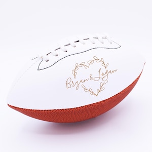 Peut inclure: Ballon de football américain blanc et marron avec les noms "Bryan & Logan" en lettres dorées dans un motif de cœur en feuilles d'or. Le ballon a des lacets et des coutures blanches.