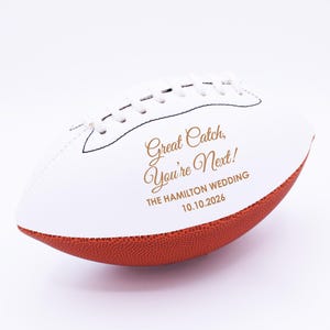 May include: White and brown football with the text "Great Catch, You're Next!" in gold script. The text "THE HAMILTON WEDDING 10.10.2026" is also printed on the ball. The football has white laces and stitching.