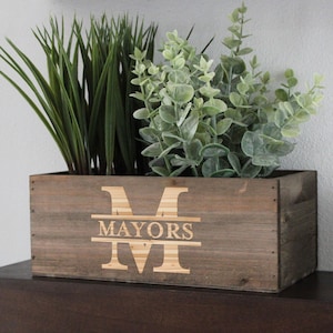 Op de afbeelding: Een houten plantenbak met de naam "MAYORS" en de initialen "M" gegraveerd op de voorkant. De plantenbak is gevuld met groene planten.