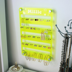 May include: Neon yellow acrylic earring holder with five rows of hooks and the name "Millie" printed on the top. The holder is mounted on a white wall and has several pairs of earrings hanging from it.