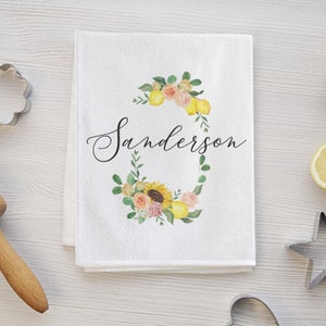 May include: White kitchen towel with the name "Sanderson" in elegant script. The towel features a watercolor floral wreath with lemons, roses, and a sunflower. Baking-themed kitchen decor.