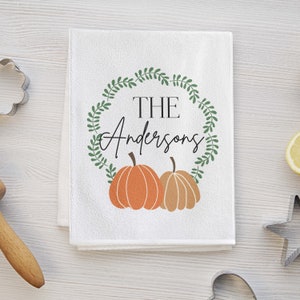 May include: White kitchen towel with a green wreath design and two orange pumpkins. The text "THE Andersons" is inside the wreath.