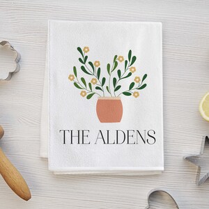 May include: White kitchen towel with a green plant with yellow flowers in a terracotta pot. The text "THE ALDENS" is printed below the plant.