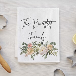 May include: White kitchen towel with the text "The Bartlett Family" in black cursive script. A floral design with pink, white, and yellow flowers and green leaves is below the text.