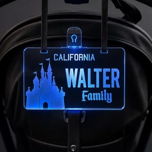 May include: A rectangular, blue-lit acrylic sign featuring a silhouette of a castle on the left, the word CALIFORNIA at the top, and the text WALTER Family in the center. The sign is suspended by black straps from a black stroller handle.
