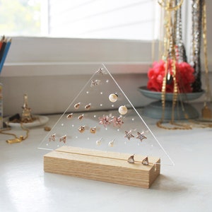 May include: A clear acrylic triangle earring holder with a wooden base. The holder has multiple holes for storing earrings. The holder is filled with various earrings, including stars, flowers, and pearls.
