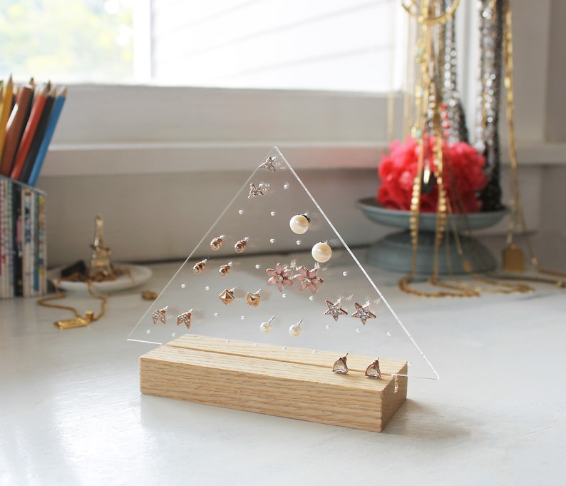 May include: A clear acrylic triangle earring holder with a wooden base. The holder has multiple holes for storing earrings. The holder is filled with various earrings, including stars, flowers, and pearls.