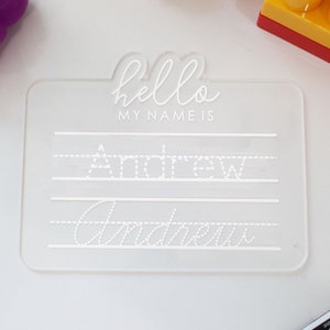 May include: A clear plastic dry-erase board with the text "hello my name is" and the name "Andrew" written on it in dotted lines. The board is designed for children to practice writing their name.