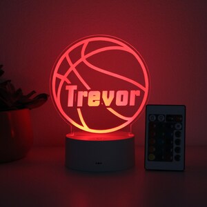 Pu&ograve; includere: Luce notturna a LED a forma di pallone da basket con il nome "Trevor" in bianco. La luce rossa &egrave; posta su una base bianca, con un telecomando. Una pianta in vaso &egrave; sullo sfondo.