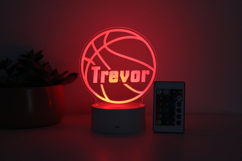 Pu&ograve; includere: Luce notturna a LED a forma di pallone da basket con il nome "Trevor" in bianco. La luce rossa &egrave; posta su una base bianca, con un telecomando. Una pianta in vaso &egrave; sullo sfondo.