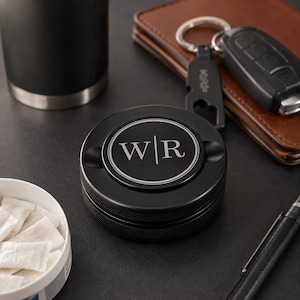 May include: A round black metal container featuring a centered circular engraving with the letters W and R separated by a vertical line. The lid has a textured knurled edge and a smooth top surface with a double-line border surrounding the monogram.
