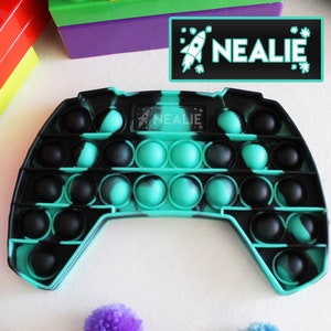 Puede incluir: Un juguete pop it fidget de color turquesa y negro con forma de mando de videojuego. El juguete tiene una pequeña etiqueta con el texto "NEALIE" en él.