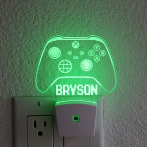 Puede incluir: Luz nocturna verde iluminada con forma de mando de videojuego con el nombre Bryson impreso debajo. La luz nocturna está enchufada a una toma de corriente estándar.