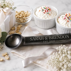 May include: A dark metallic ice cream scoop featuring the engraved text SARAH & BRENDON and EST. 2020 on the handle. The scoop is positioned on a light-colored fabric napkin, surrounded by white flowers, a gift box, and bowls of ice cream.