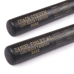 May include: Two black baseball bats with engraved text. One bat reads "COACH STEVEN LONG BEACH YANKEES 2022" and lists names. The other bat reads "DANIEL FINLEY #1 LONG BEACH YANKEES 2022" and lists names. The bats are made of wood.